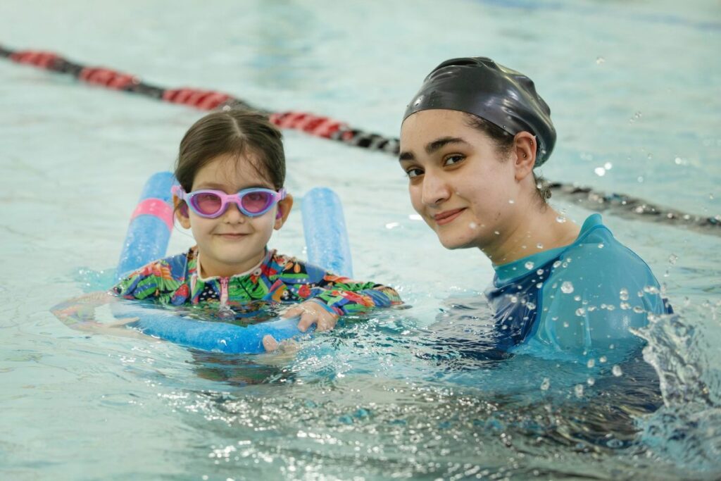 Swim teacher smiling beside a young girl in pink goggles who is holding a kickboard and supported by a pool noodle